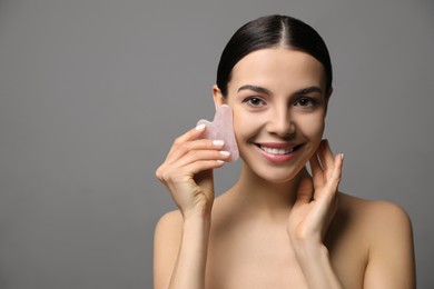 Beautiful young woman doing facial massage with gua sha tool on grey background. Space for text