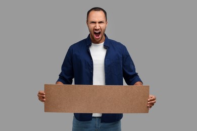 Photo of Angry man holding blank cardboard banner on grey background, space for text