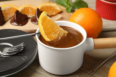 Photo of Dipping slice of orange into fondue pot with milk chocolate on wooden table, closeup