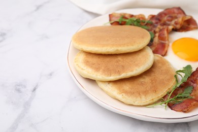 Photo of Plate with tasty pancakes, fried egg, arugula and bacon on white marble table, closeup. Space for text