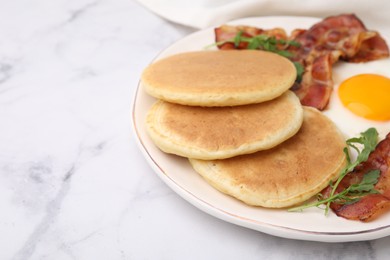 Plate with tasty pancakes, fried egg, arugula and bacon on white marble table, closeup. Space for text