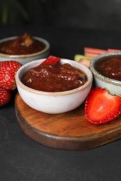Tasty rhubarb jam in bowls and strawberries on dark table
