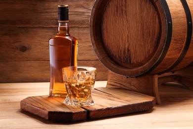 Whiskey with ice cubes in glass, bottle and barrel on wooden table, space for text