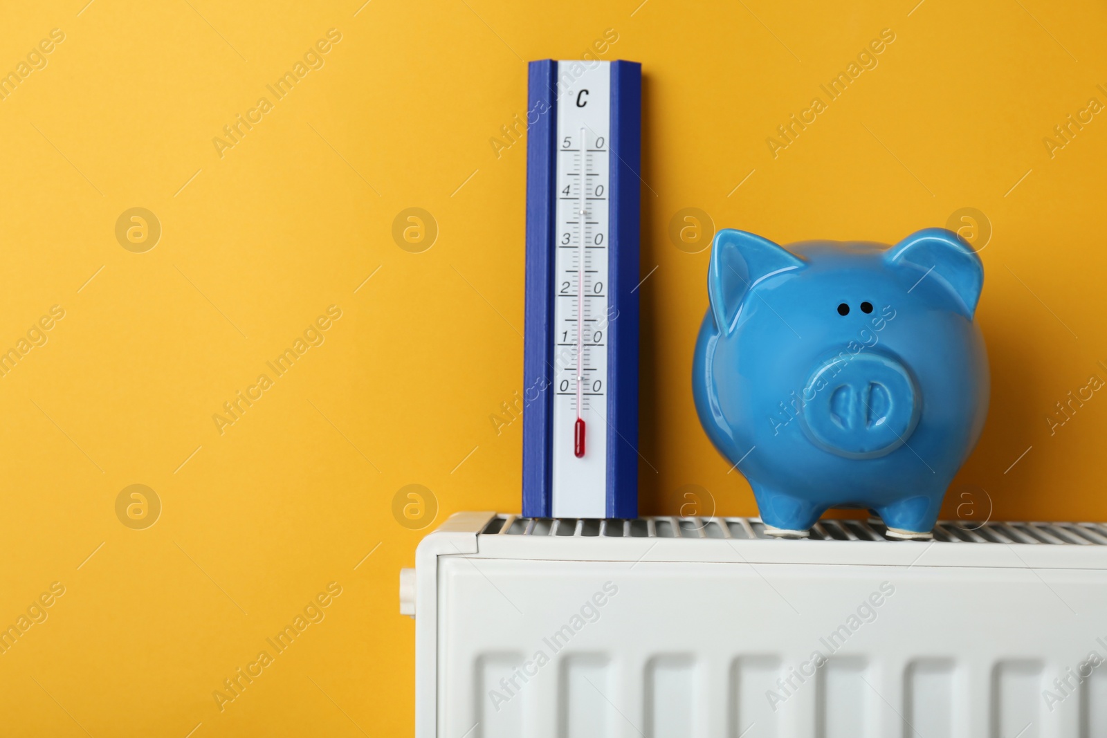 Photo of Piggy bank and thermometer on heating radiator against orange background, space for text