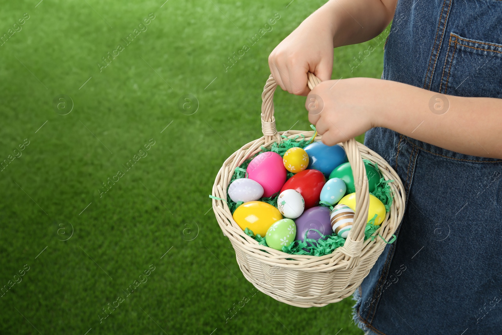 Photo of Little girl with basket full of Easter eggs on green grass, closeup. Space for text