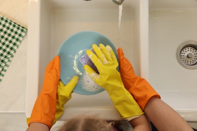 Photo of Mother and daughter in protective gloves washing plate above sink, top view