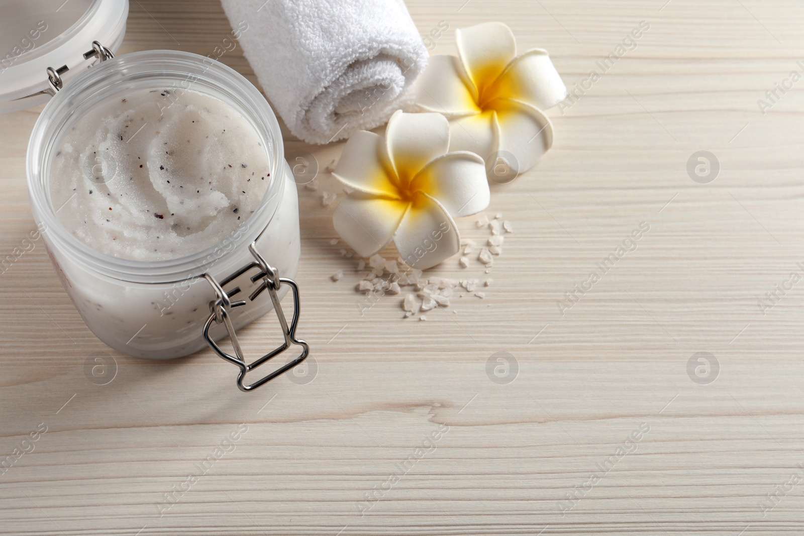 Photo of Body scrub in glass jar, towel and plumeria flowers on white wooden table, above view. Space for text