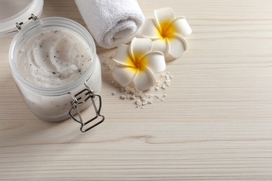 Body scrub in glass jar, towel and plumeria flowers on white wooden table, above view. Space for text