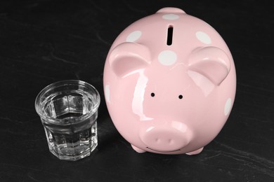 Photo of Water scarcity concept. Piggy bank and glass of drink on dark table
