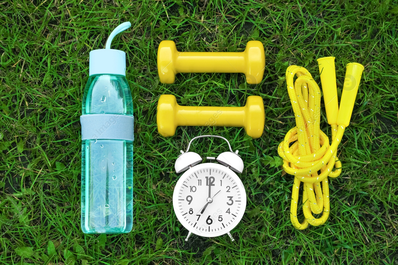 Photo of Alarm clock, skipping rope, dumbbells and bottle of water on green grass, flat lay. Morning exercise