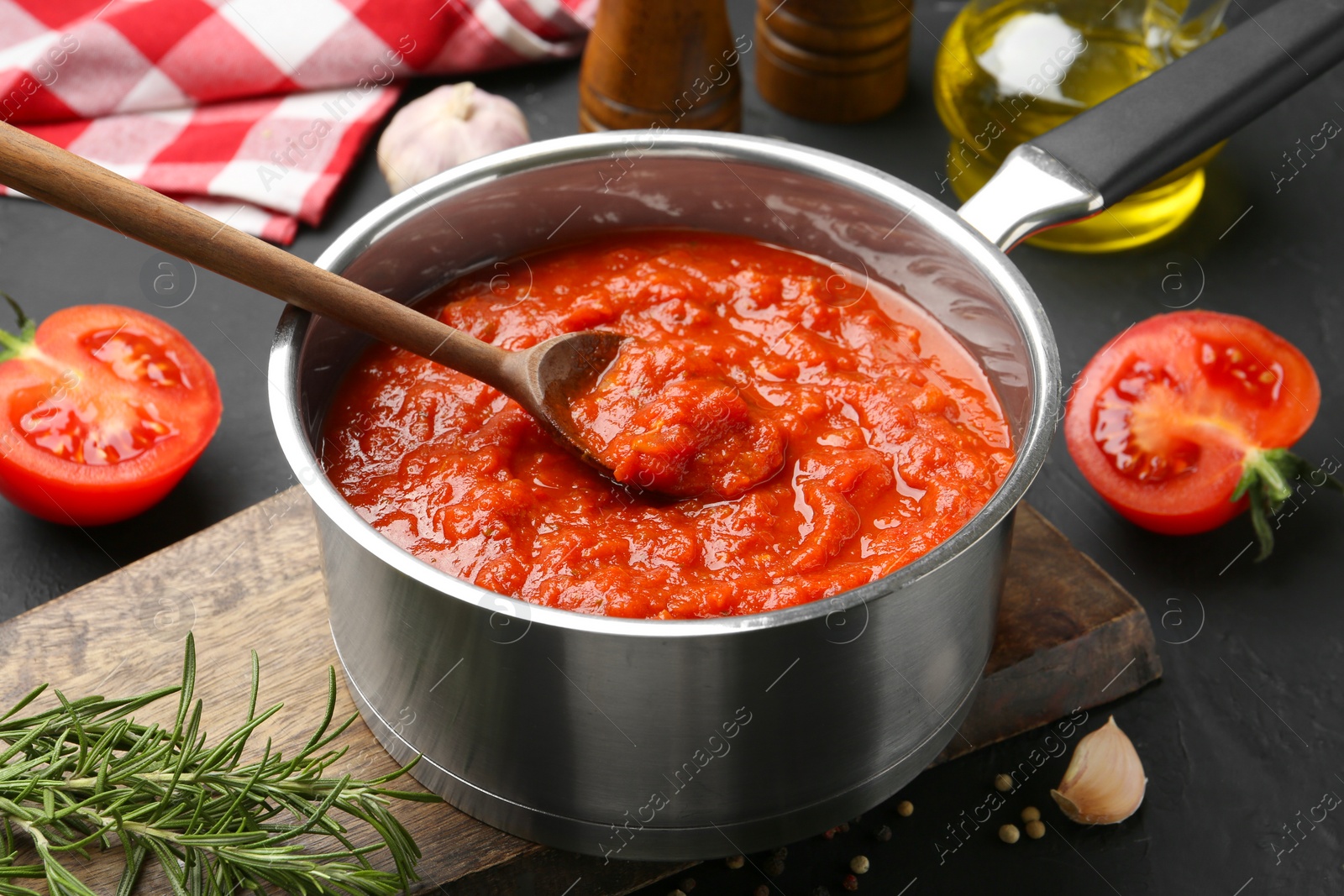 Photo of Homemade tomato sauce in pot, spoon and fresh ingredients on dark table