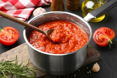 Photo of Homemade tomato sauce in pot, spoon and fresh ingredients on dark table
