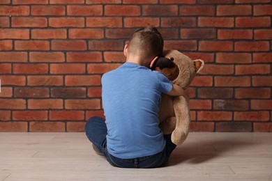 Photo of Child abuse. Upset boy with teddy bear sitting on floor near brick wall indoors, back view
