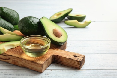 Photo of Board with bowl of natural oil and avocados on white wooden background. Space for text