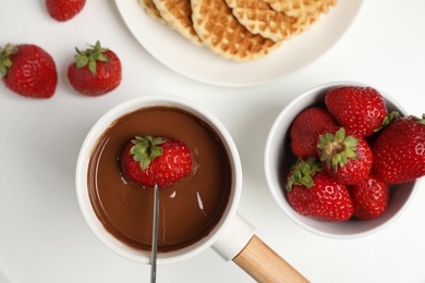 Photo of Dipping strawberry into pot with chocolate fondue on white table, flat lay