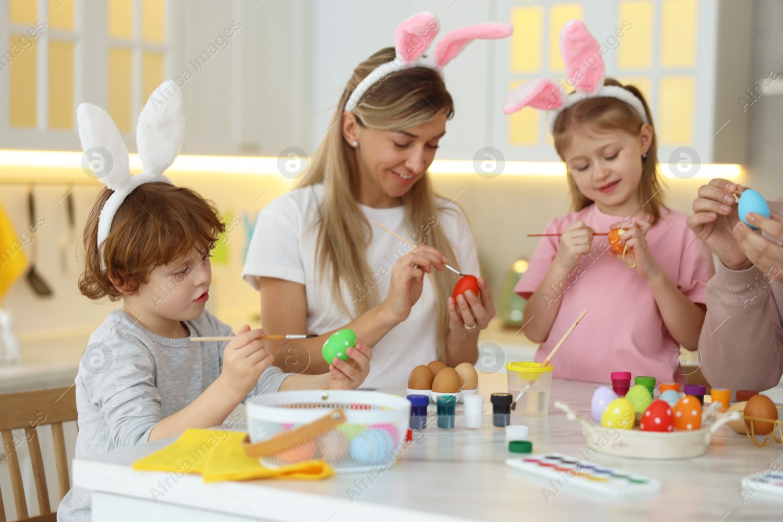 Photo of Easter celebration. Happy family with bunny ears painting eggs at white marble table in kitchen