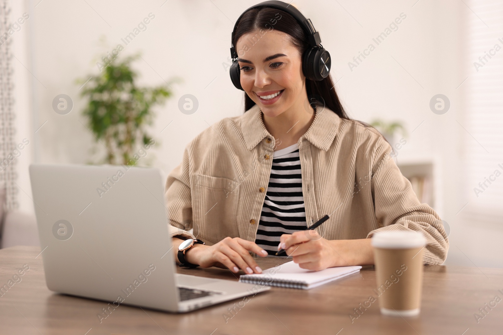 Photo of Young woman in headphones watching webinar at table in room