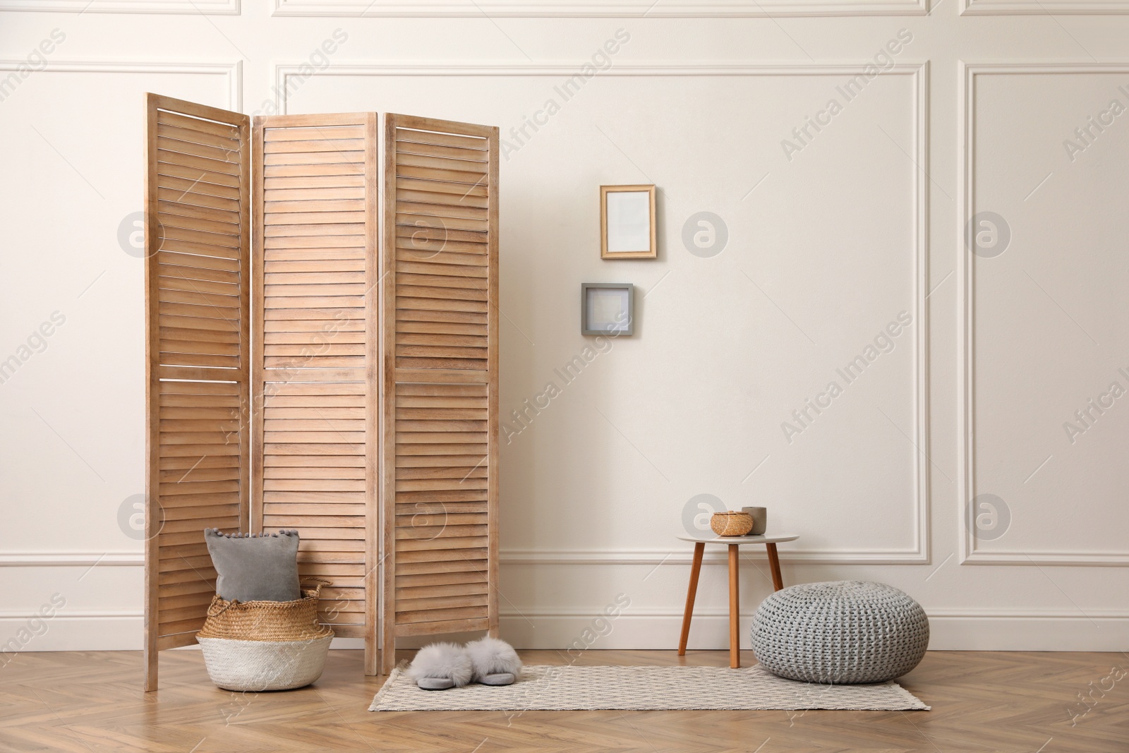 Photo of Folding screen, small table and knitted pouf near white wall in room. Interior design