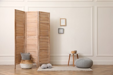 Folding screen, small table and knitted pouf near white wall in room. Interior design