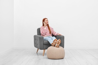 Happy young woman talking on smartphone while sitting in armchair near white wall indoors