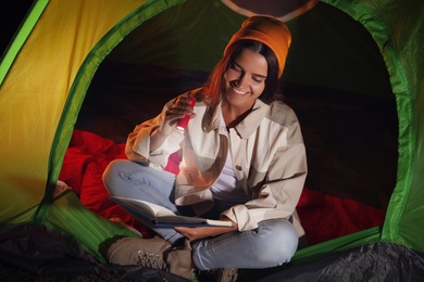 Photo of Young woman with flashlight reading book in tent at night