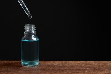 Dripping cosmetic serum from pipette into bottle at wooden table against black background, space for text