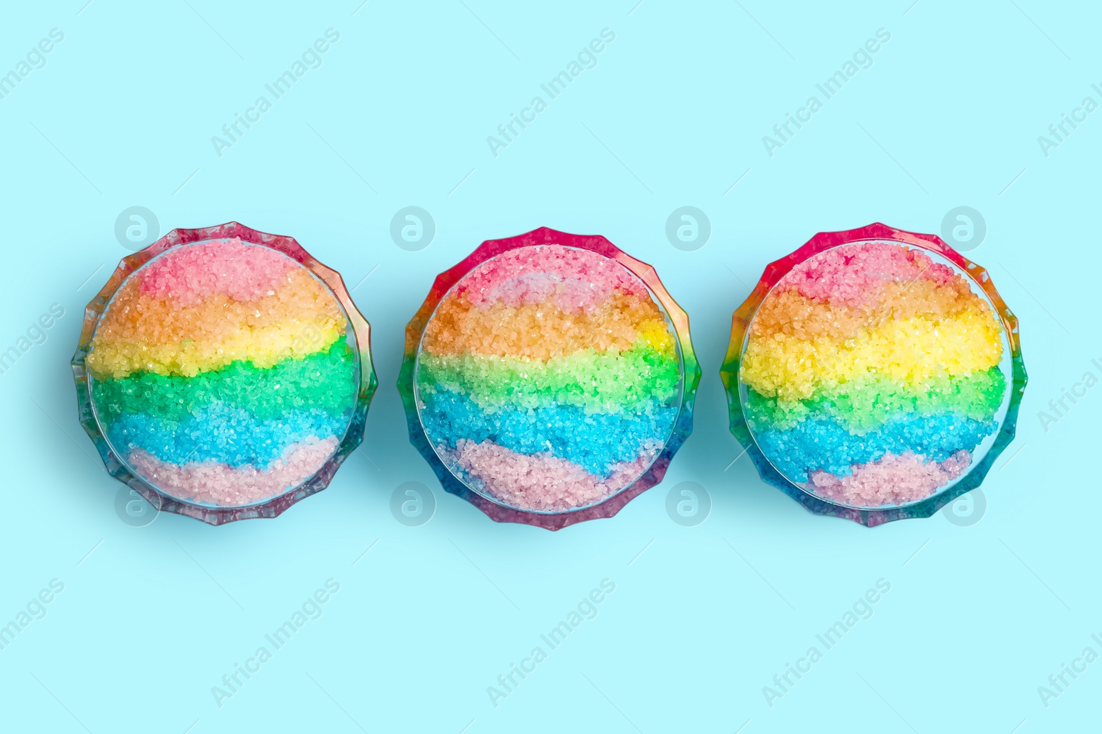 Photo of Rainbow shaving ice in glass dessert bowls on light blue background, flat lay