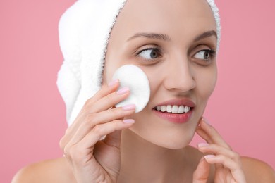 Young woman cleaning her face with cotton pad on pink background, closeup