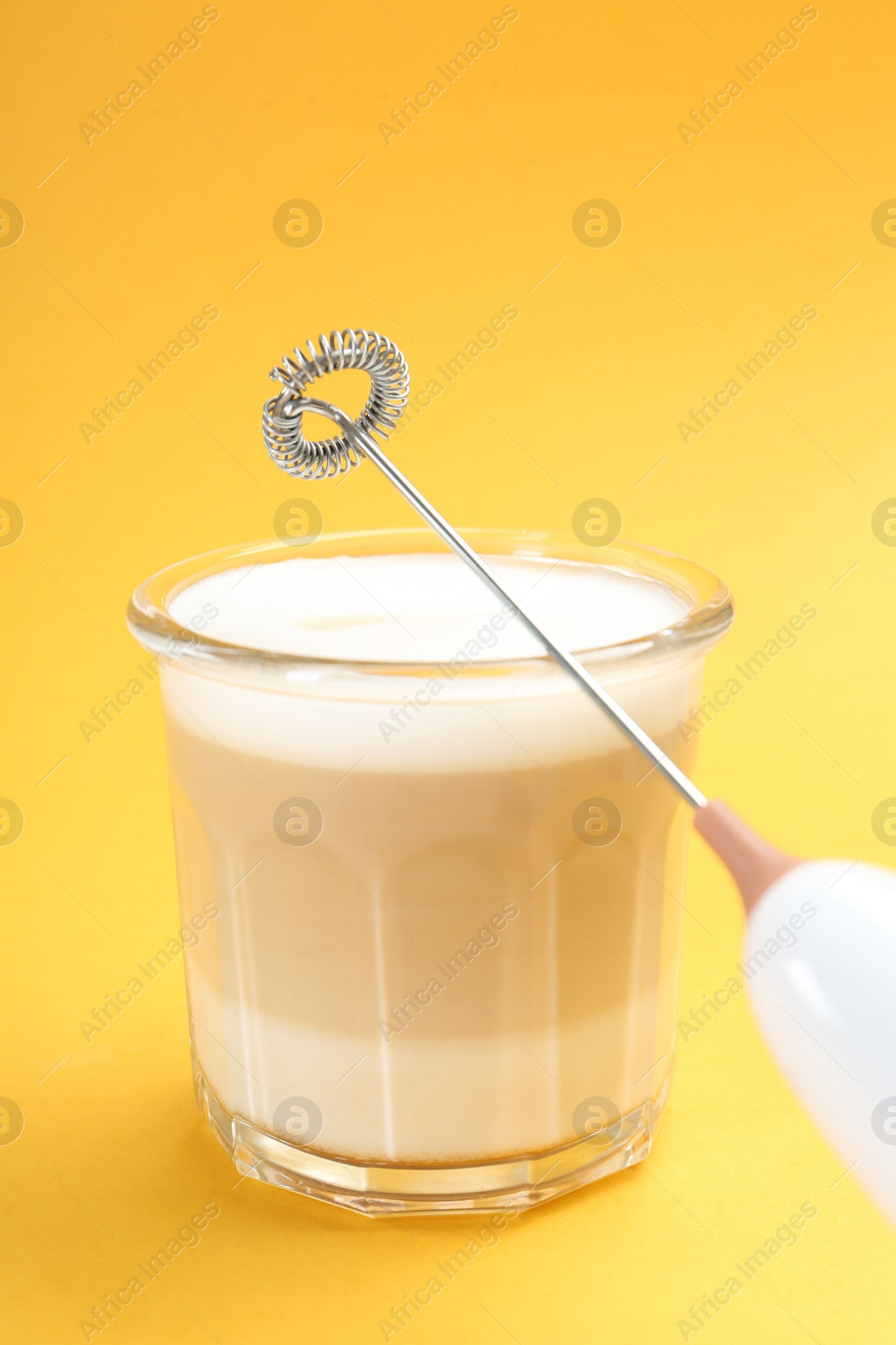 Photo of Mini mixer (milk frother) and tasty cappuccino in glass on yellow background