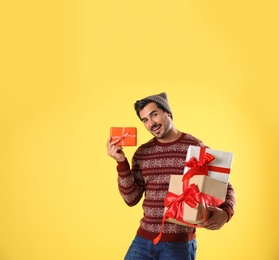 Happy man in Christmas sweater and hat holding gift boxes on yellow background