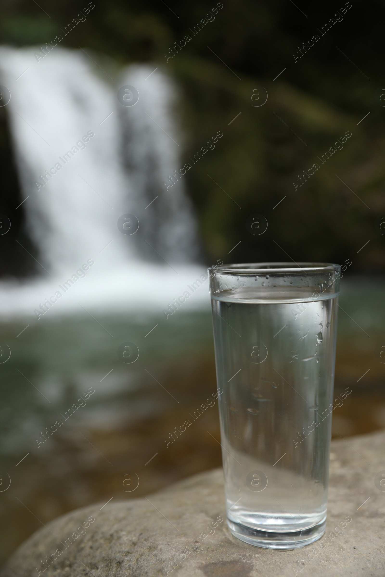 Photo of Glass of water on stone near waterfall outdoors. Space for text