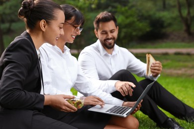 Happy colleagues with laptop having business lunch on green grass outdoors, selective focus