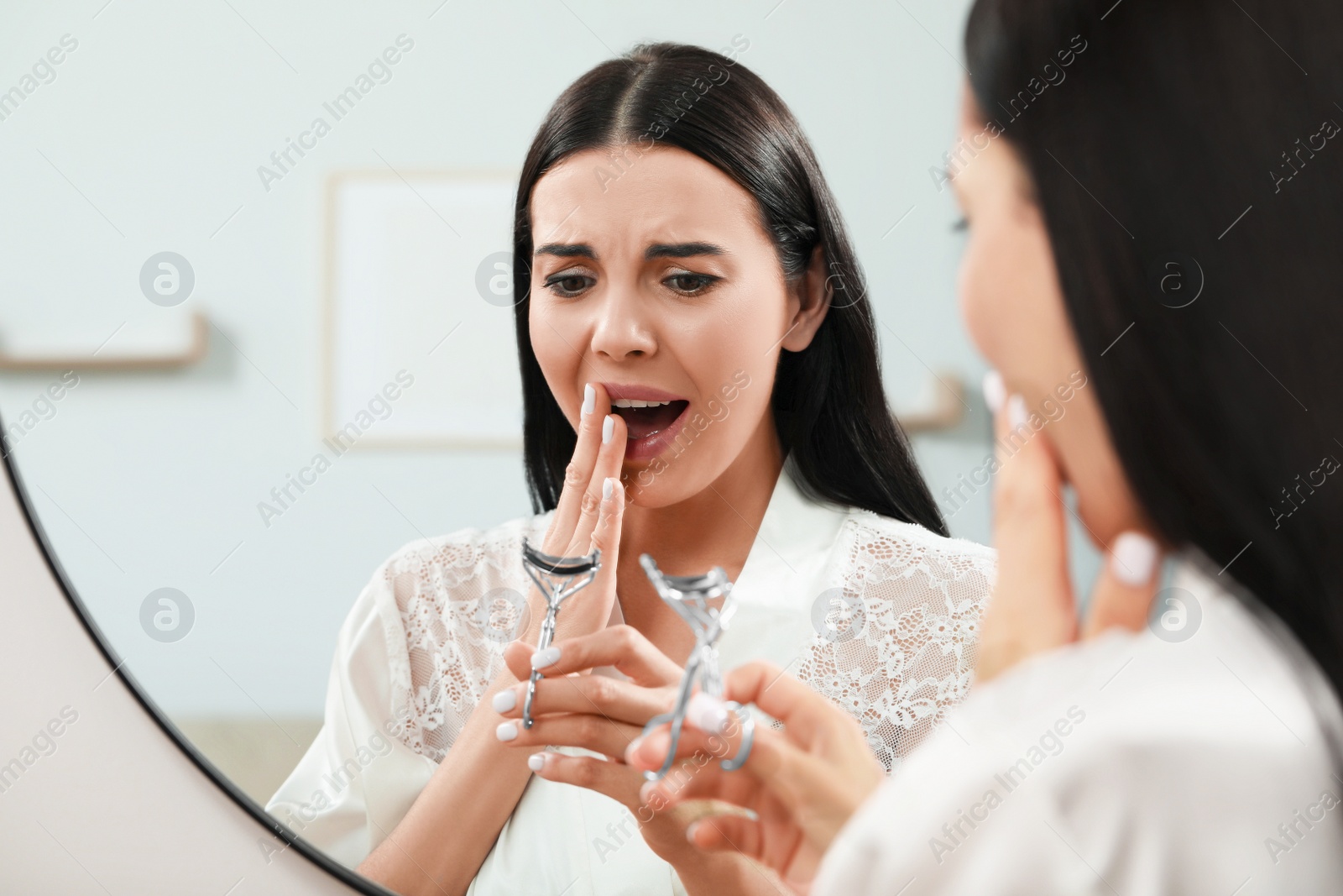 Photo of Emotional young woman with eyelash curler near mirror indoors