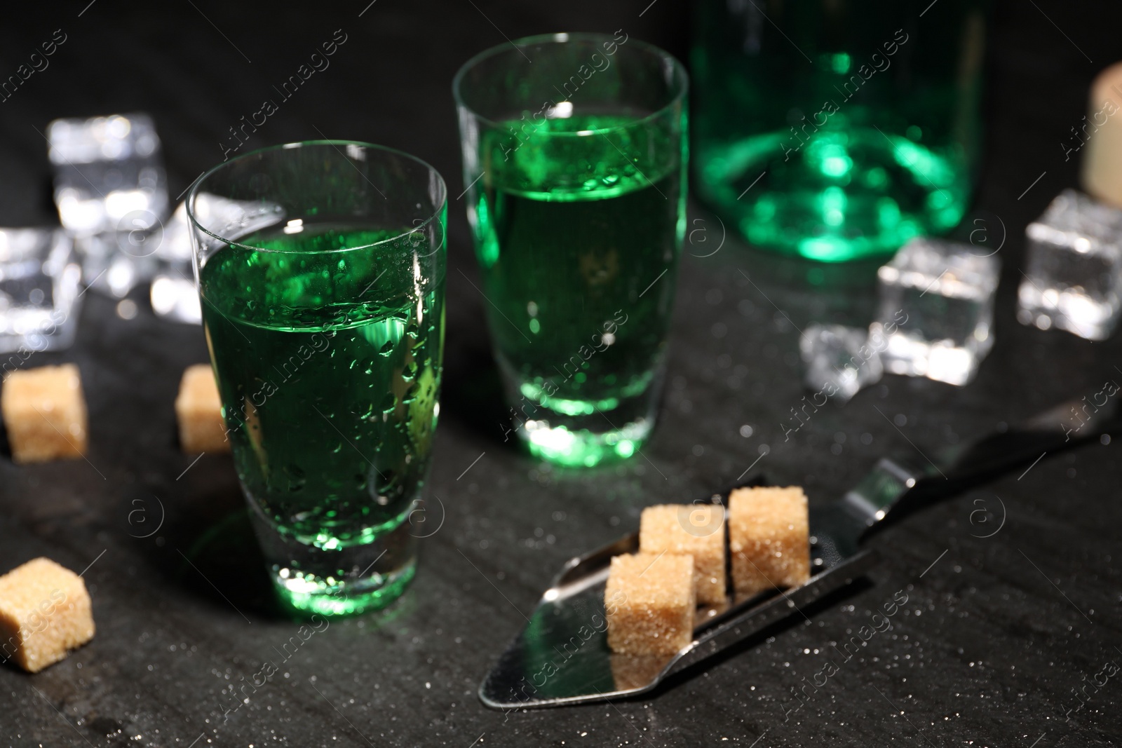 Photo of Absinthe in shot glasses, spoon, brown sugar and ice cubes on gray table, closeup. Alcoholic drink