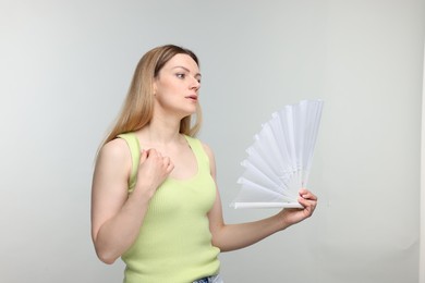 Beautiful woman waving white hand fan to cool herself on light grey background