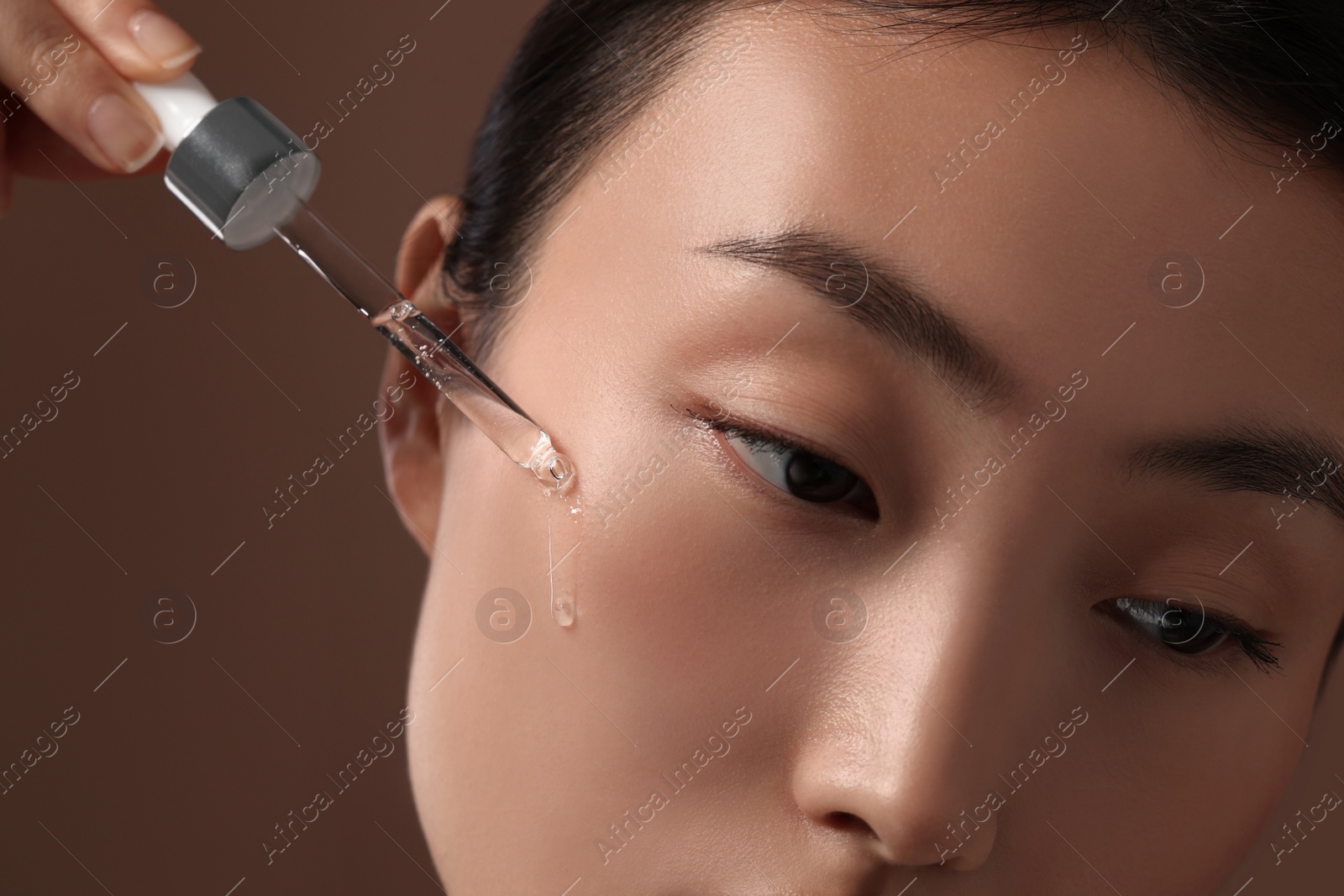 Photo of Beautiful young woman applying cosmetic serum onto her face on brown background, closeup