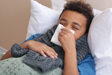 Photo of African-American boy with scarf and tissue blowing nose in bed indoors. Cold symptoms