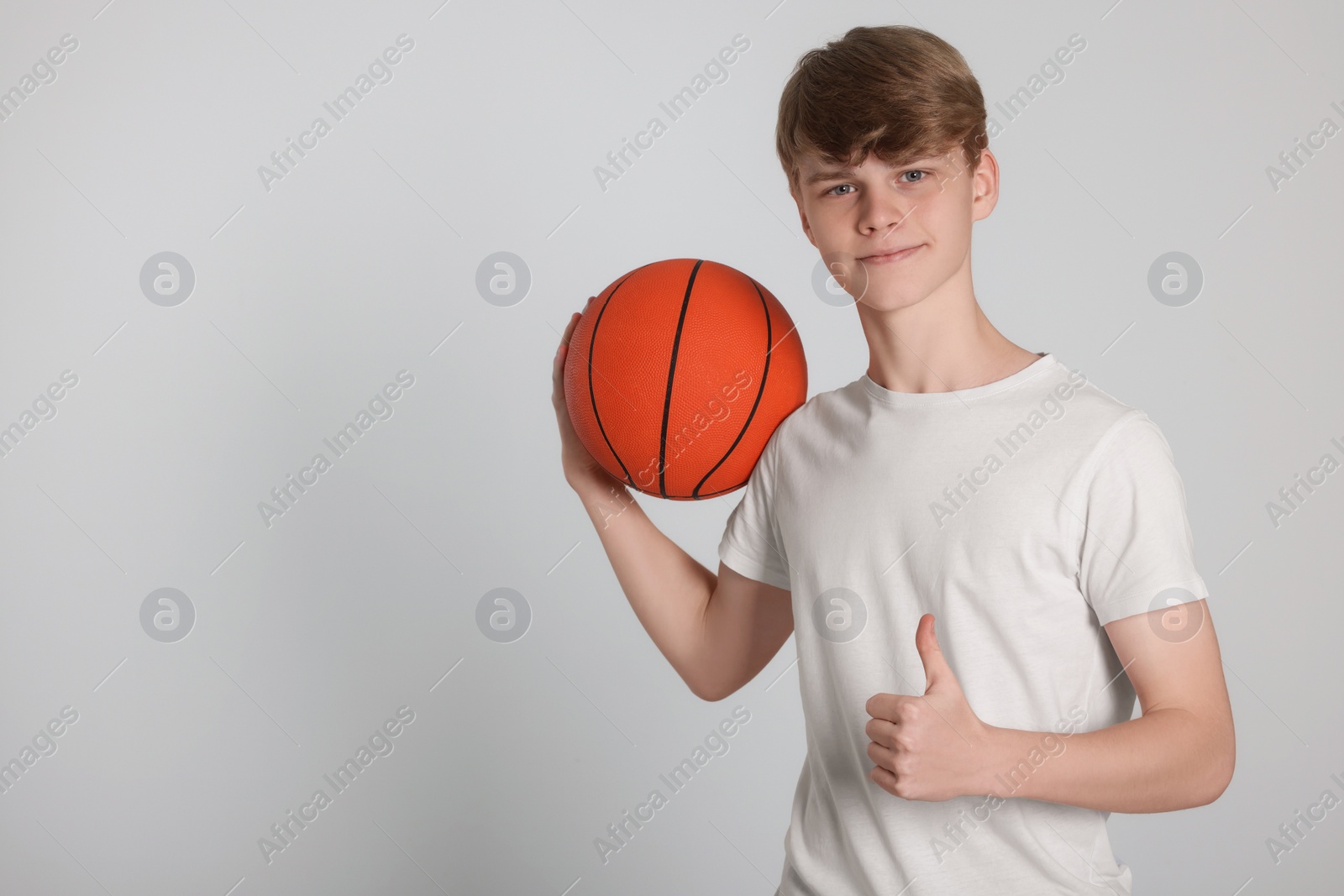 Photo of Teenage boy with basketball ball showing thumbs up on light grey background. Space for text