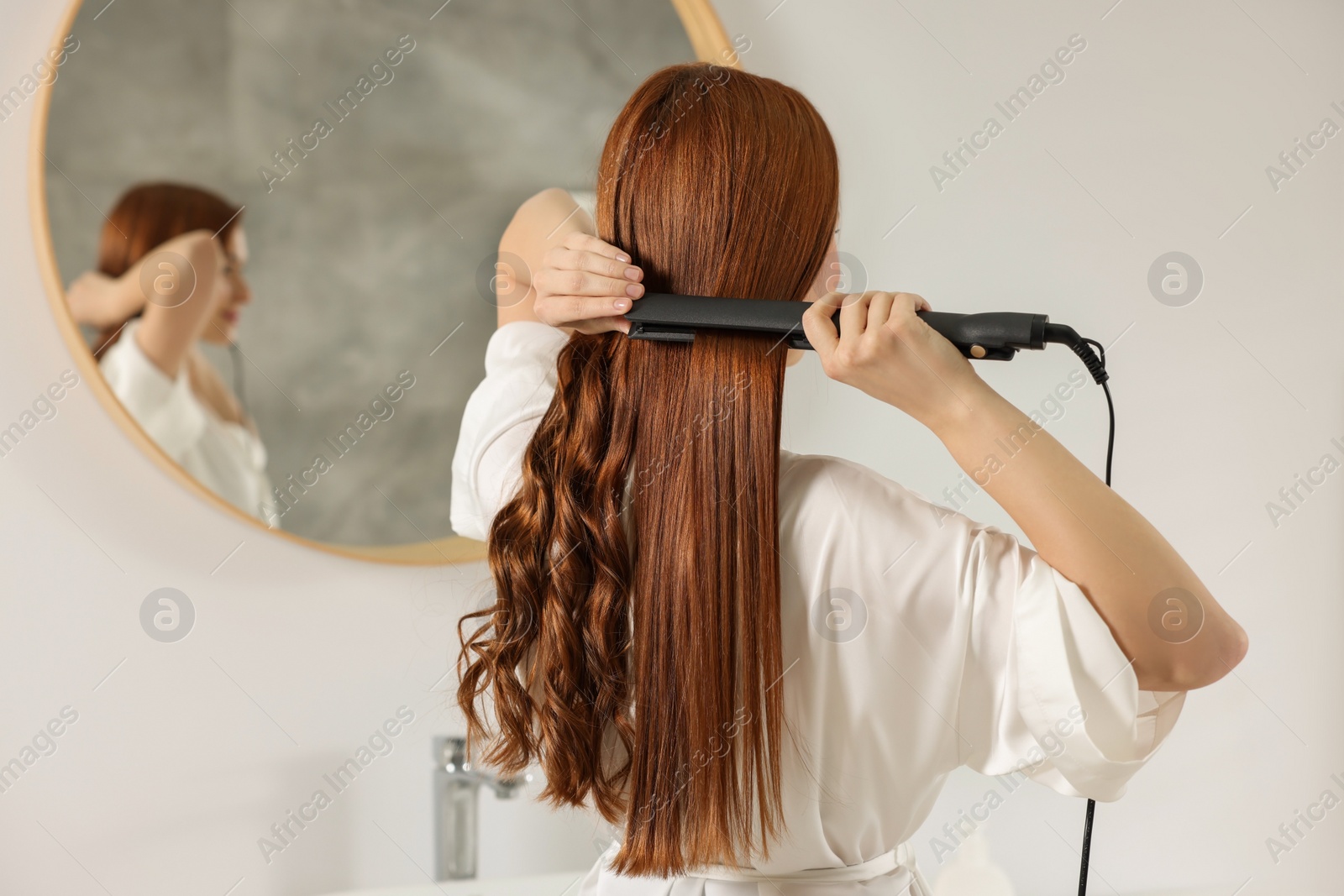 Photo of Beautiful woman using hair iron in room, back view. Space for text