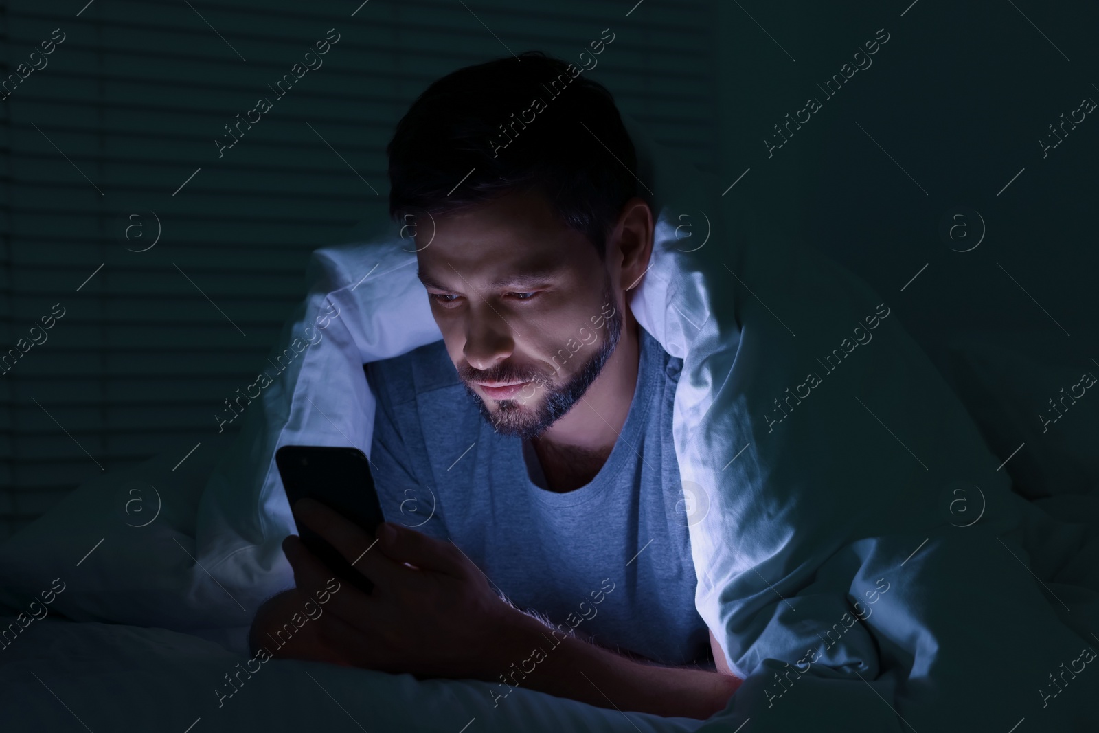 Photo of Man using smartphone under blanket in bed at night. Internet addiction