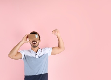 Young man using cardboard virtual reality headset on color background. Space for text