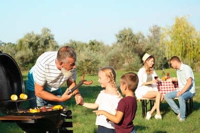 Photo of Grandfather with little kids cooking food on barbecue grill and their family in park