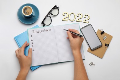 Woman filling list of resolutions for 2022 new year in notebook on white background, top view