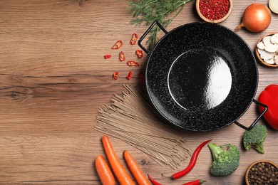 Photo of Empty iron wok surrounded by raw ingredients on wooden table, flat lay. Space for text