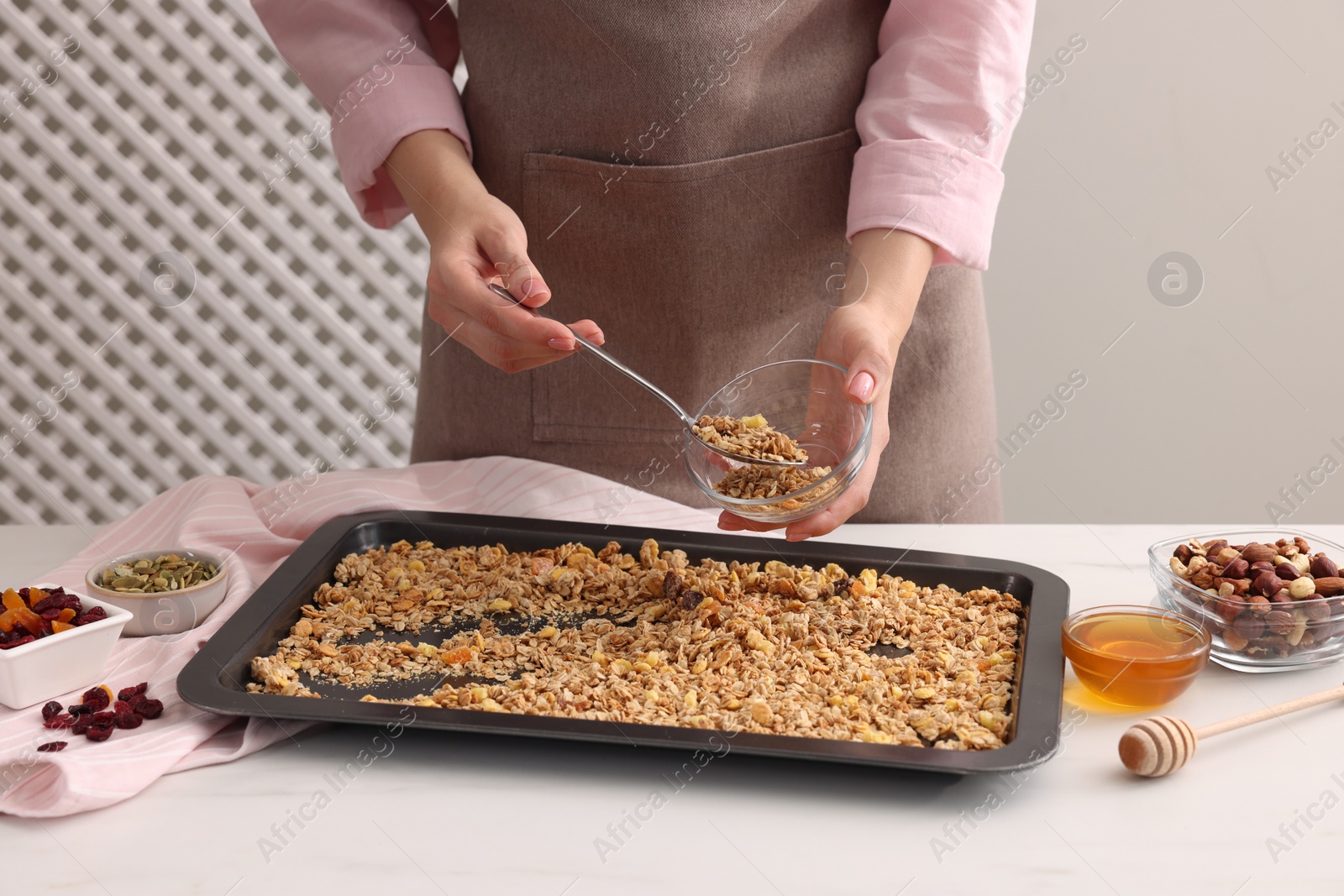 Photo of Woman putting granola from baking tray into glass bowl at white table, closeup