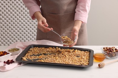 Woman putting granola from baking tray into glass bowl at white table, closeup