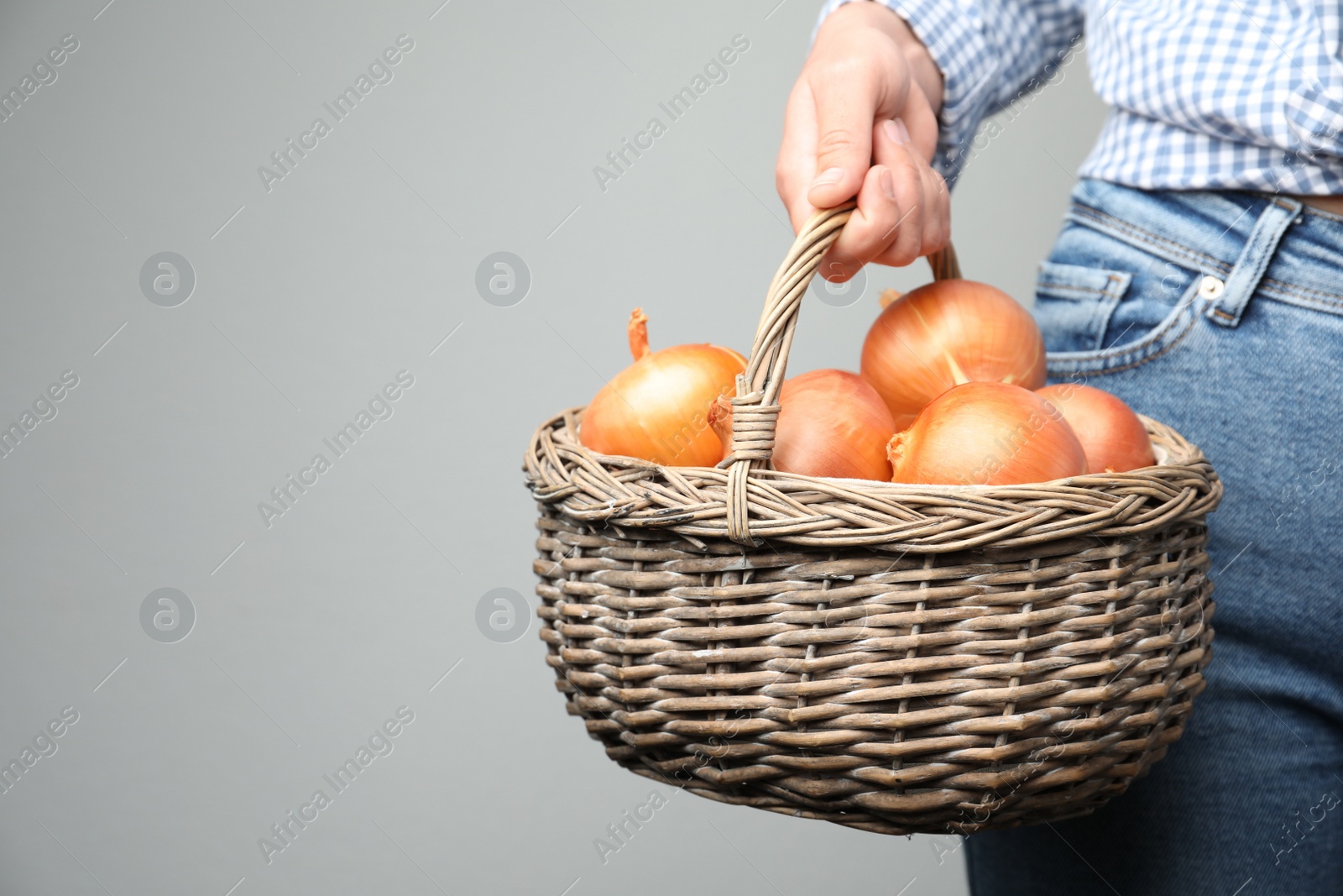 Photo of Woman holding wicker basket with raw yellow onion bulbs on grey background, closeup. Space for text