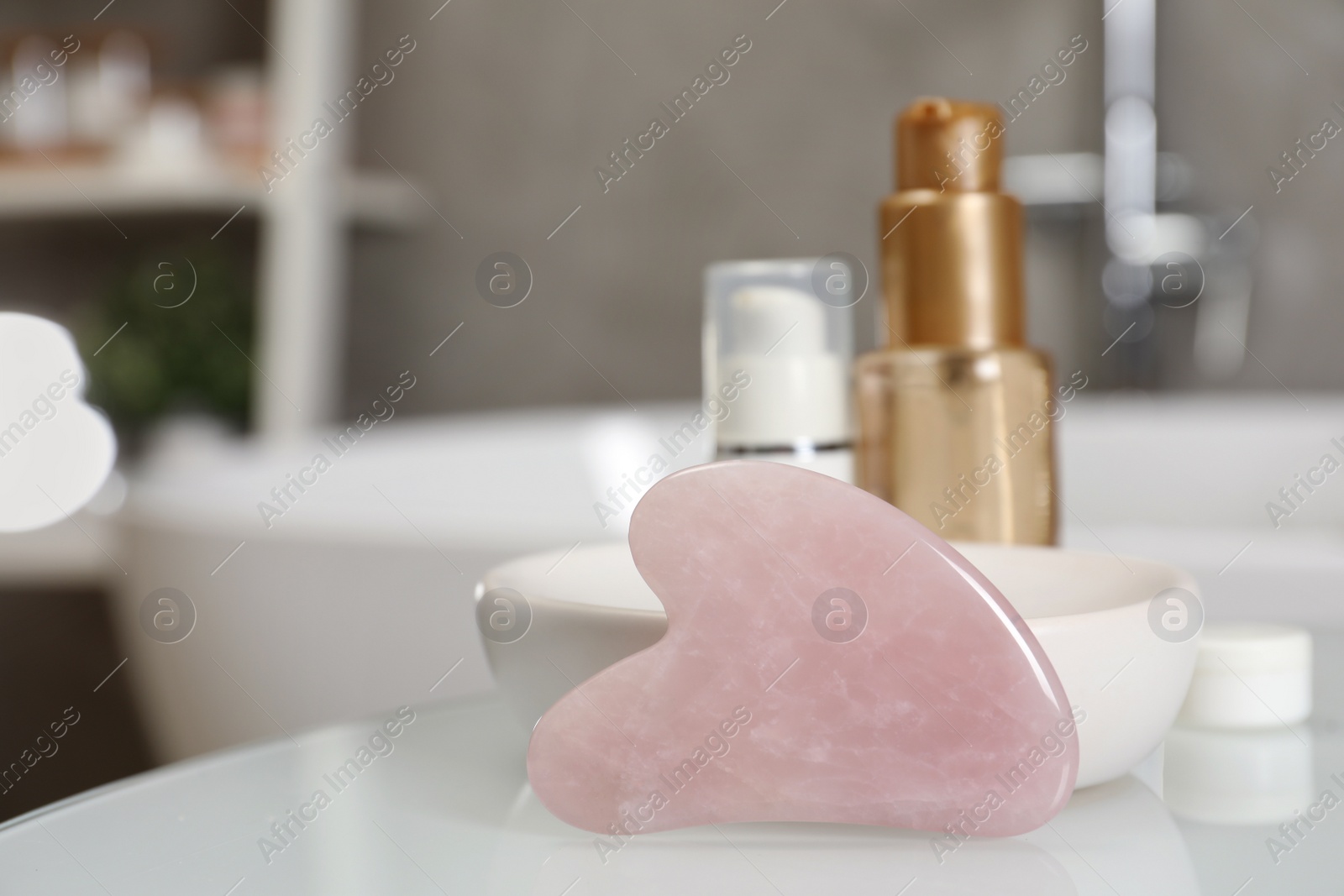 Photo of Rose quartz gua sha tool and cosmetic products on white table in bathroom. Space for text