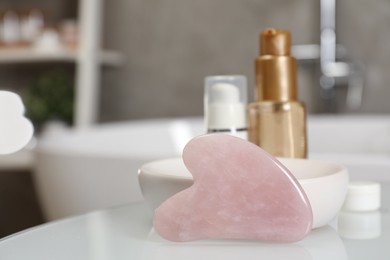 Rose quartz gua sha tool and cosmetic products on white table in bathroom. Space for text