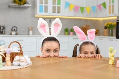 Photo of Mother with daughter wearing bunny ears headbands and peeking over table in kitchen. Easter celebration